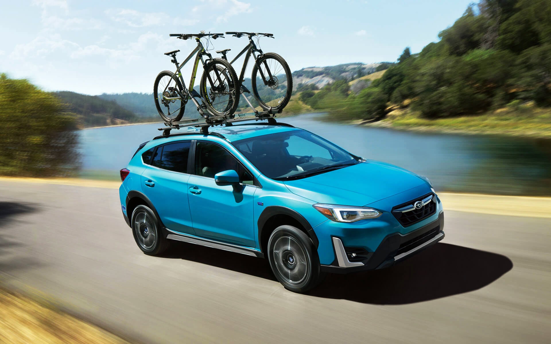 A blue Crosstrek Hybrid with two bicycles on its roof rack driving beside a river | Subaru World of Hackettstown in Hackettstown NJ